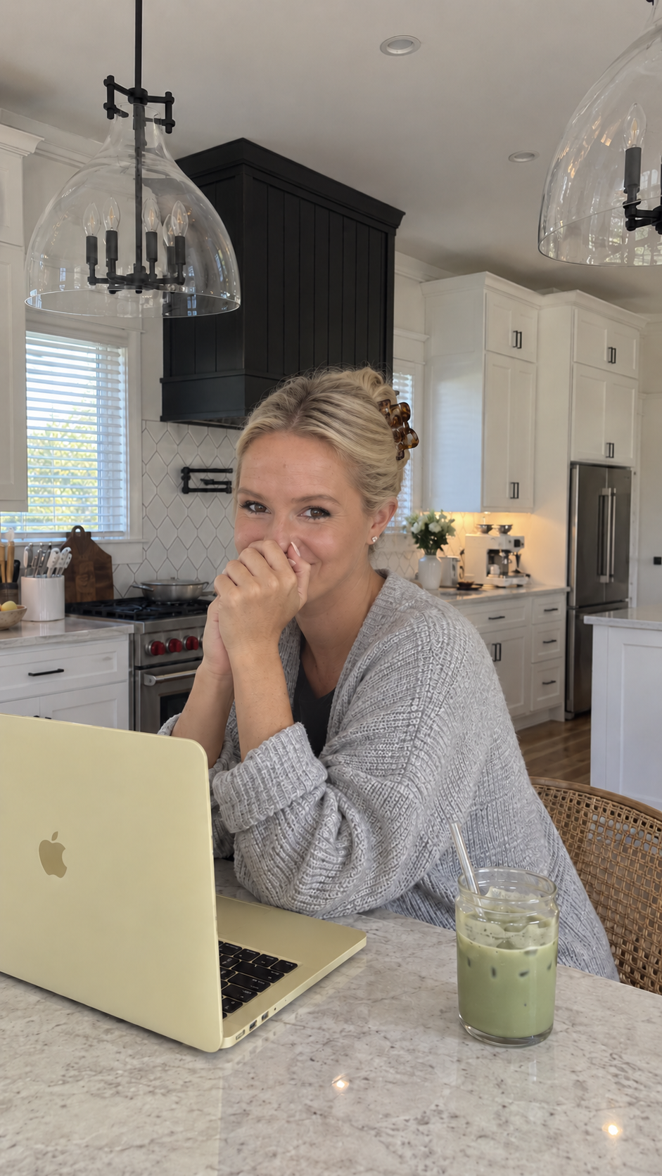 Katie Cross at her kitchen island in a gray sweater with an iced matcha and her gold laptop